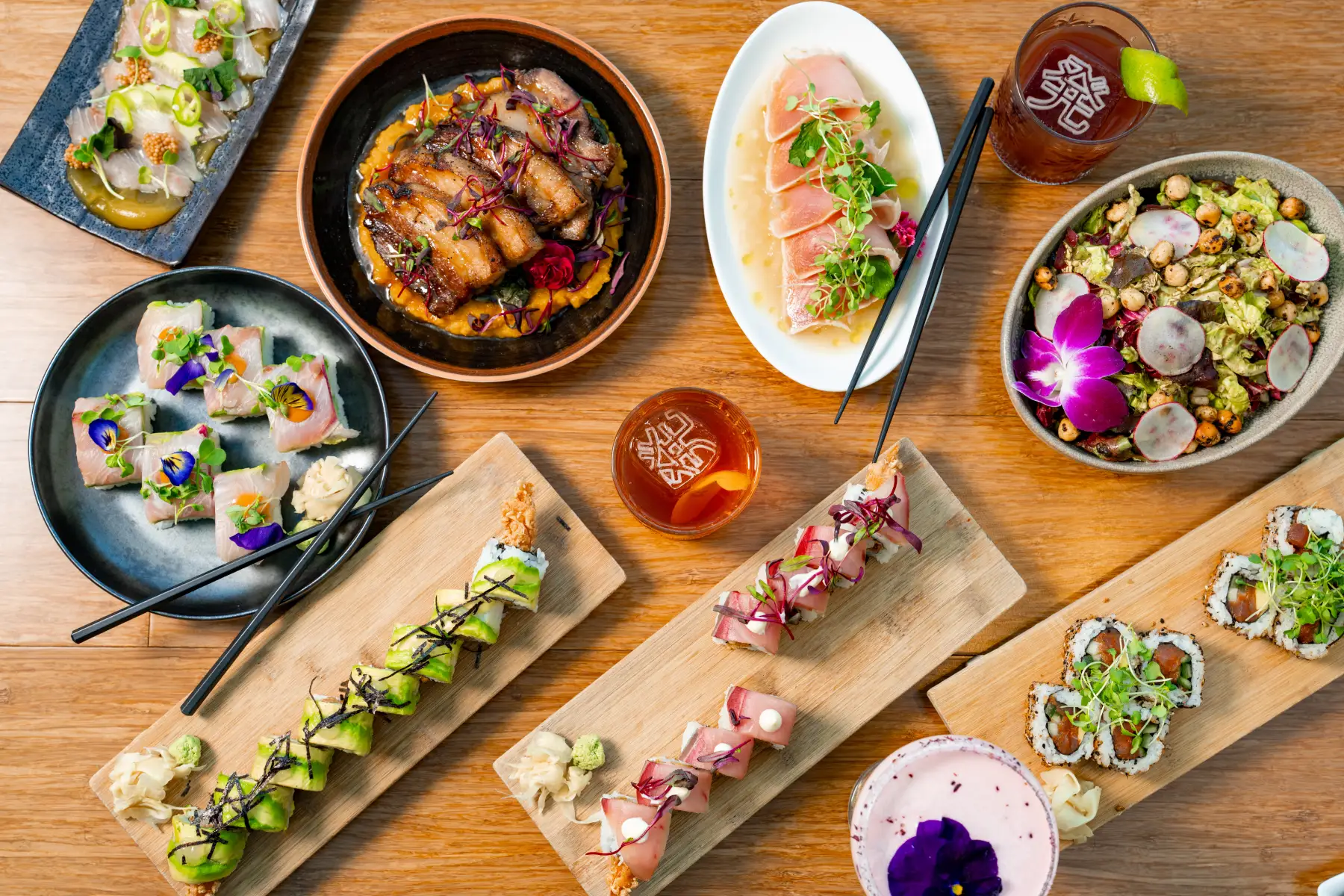 Top-down view of a diverse sushi and Asian fusion spread on a wooden table, featuring rolls, sashimi, and cocktails.