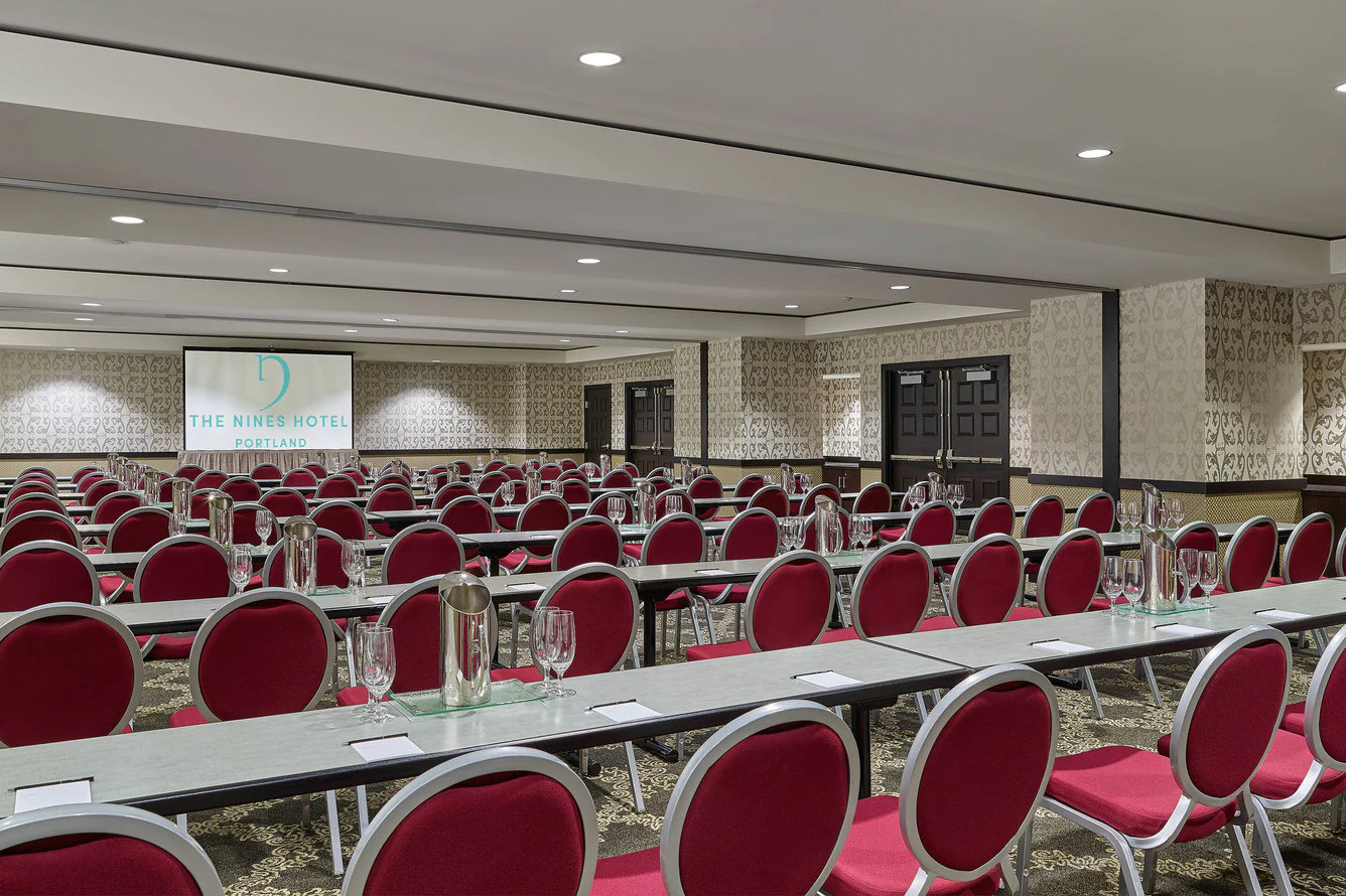 Conference room at The Nines Hotel Portland set up with rows of tables, red chairs, and a presentation screen.