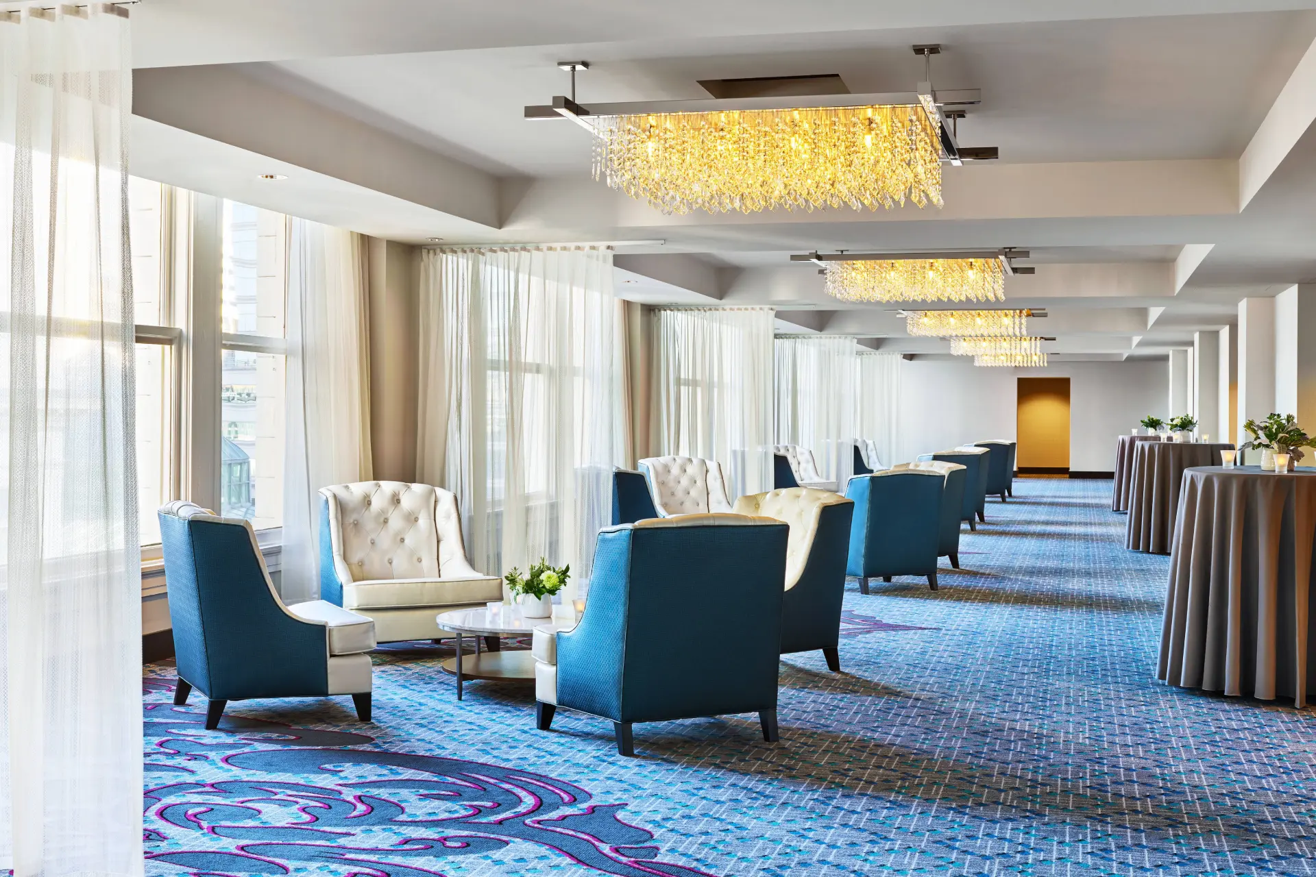Elegant hotel lounge at The Nines featuring blue and cream armchairs, crystal chandeliers, and patterned carpet.