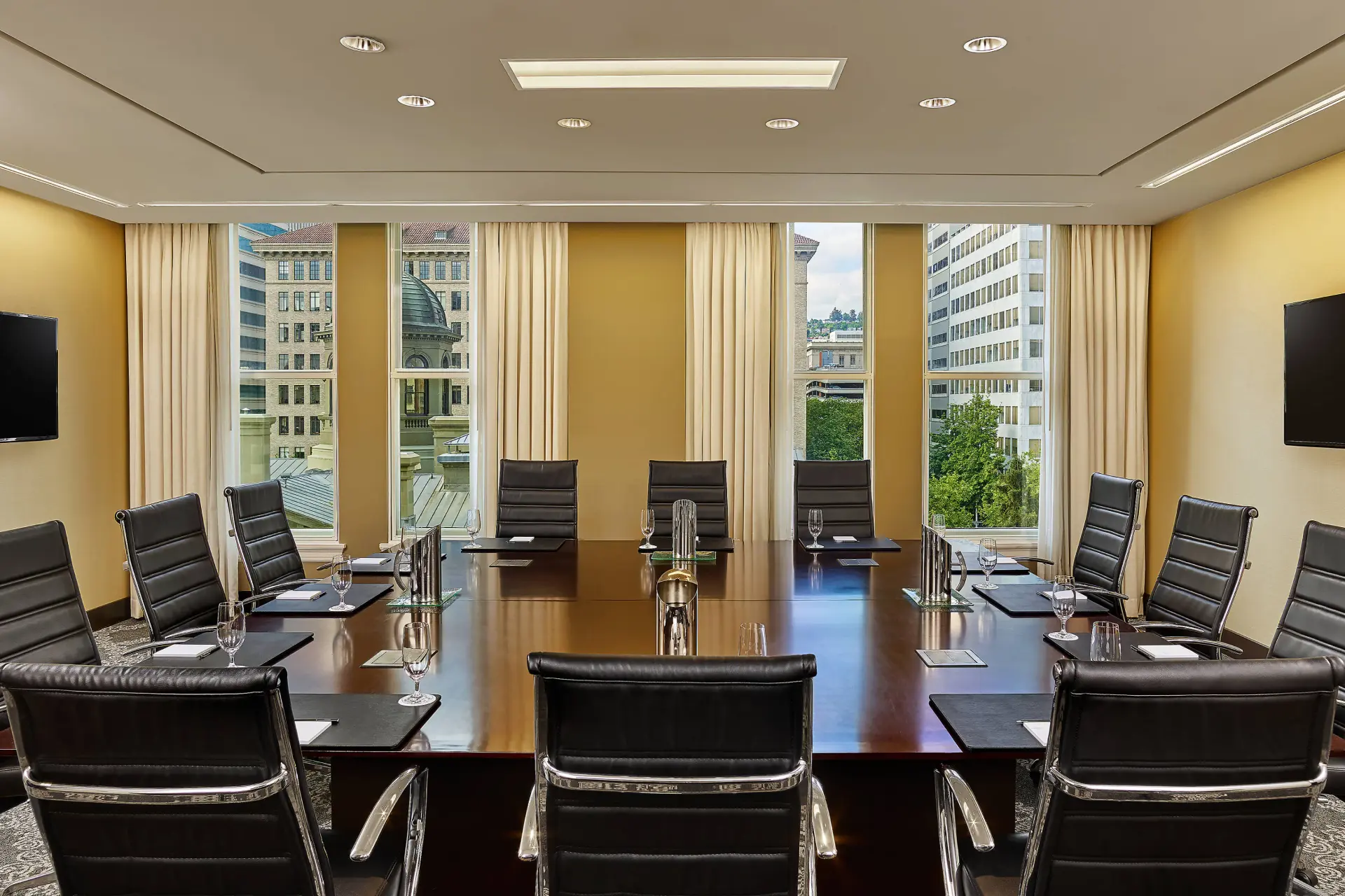 Spacious conference room with a large polished wooden table, black leather chairs, and city views through tall windows.