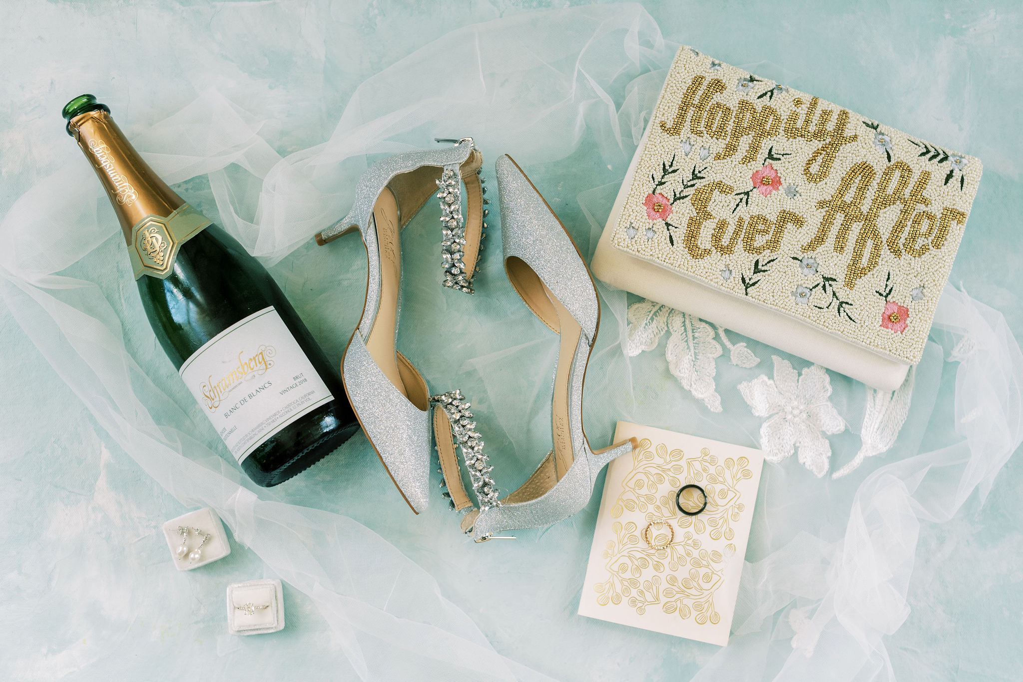 Bridal accessories including champagne, silver heels, and rings on tulle