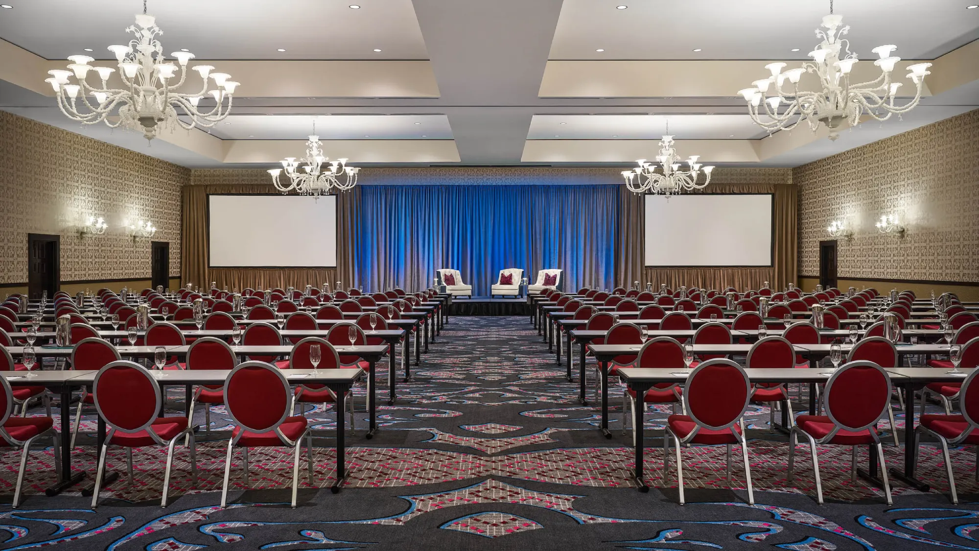Spacious conference room with rows of tables, red chairs, and chandeliers facing a stage with blue curtains.