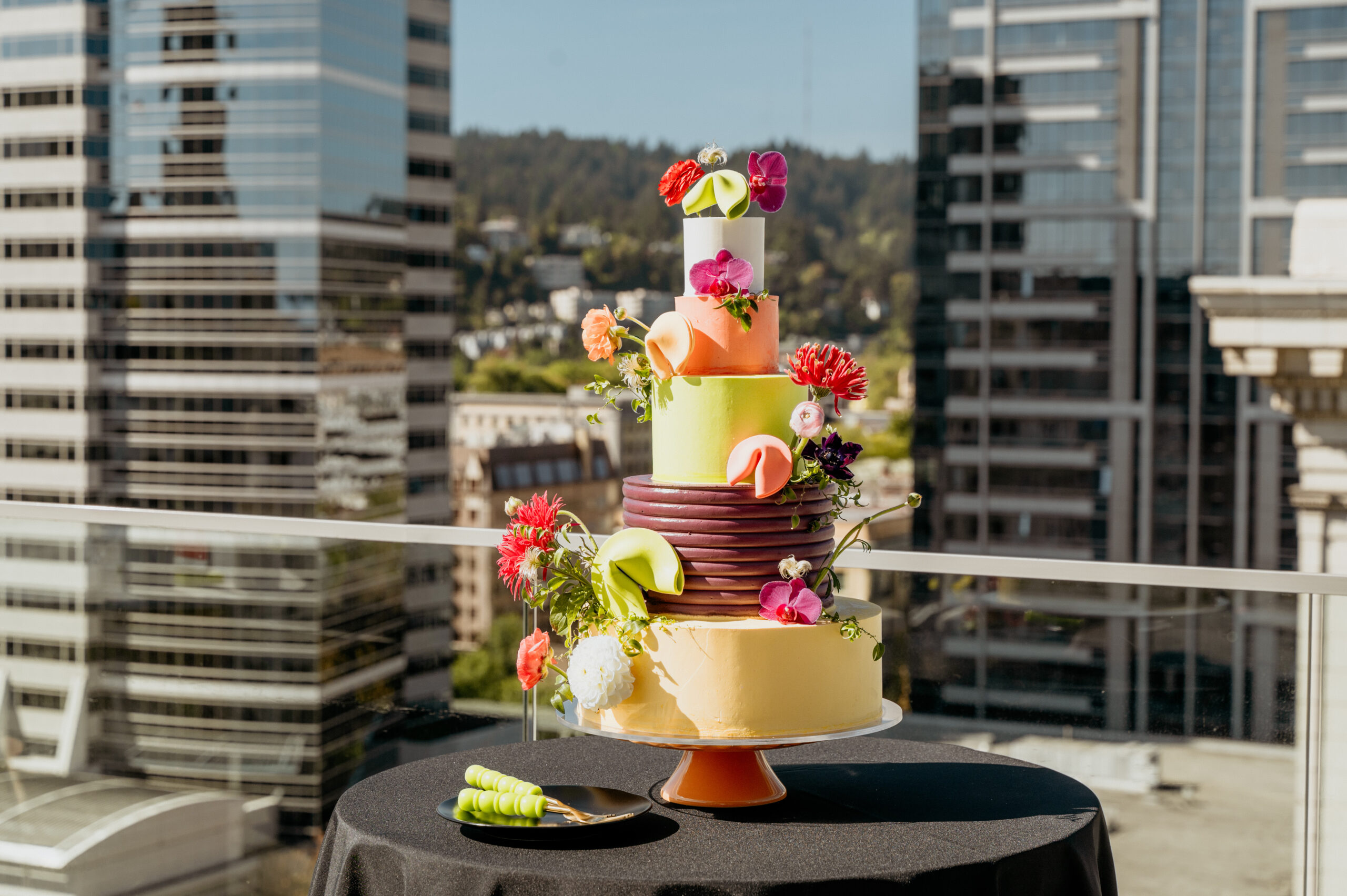Colorful multi-tiered wedding cake with floral decorations on a balcony overlooking a city skyline.