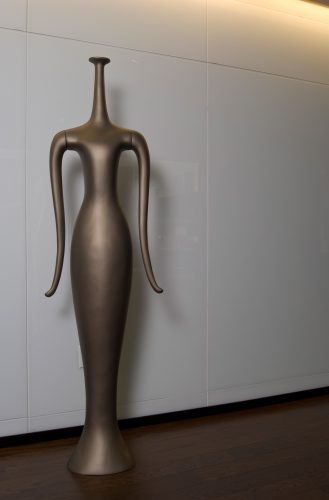 Tall abstract bronze sculpture of a stylized female figure with elongated arms against a white wall.