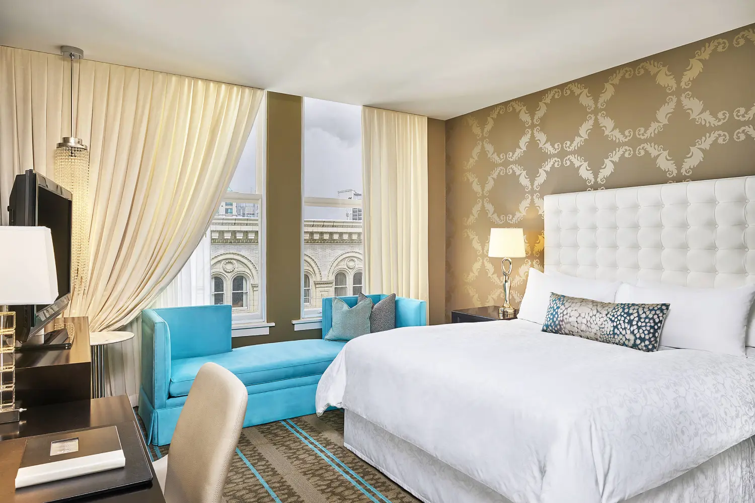 Luxury hotel room with white bed, blue chaise lounge, and patterned wallpaper.