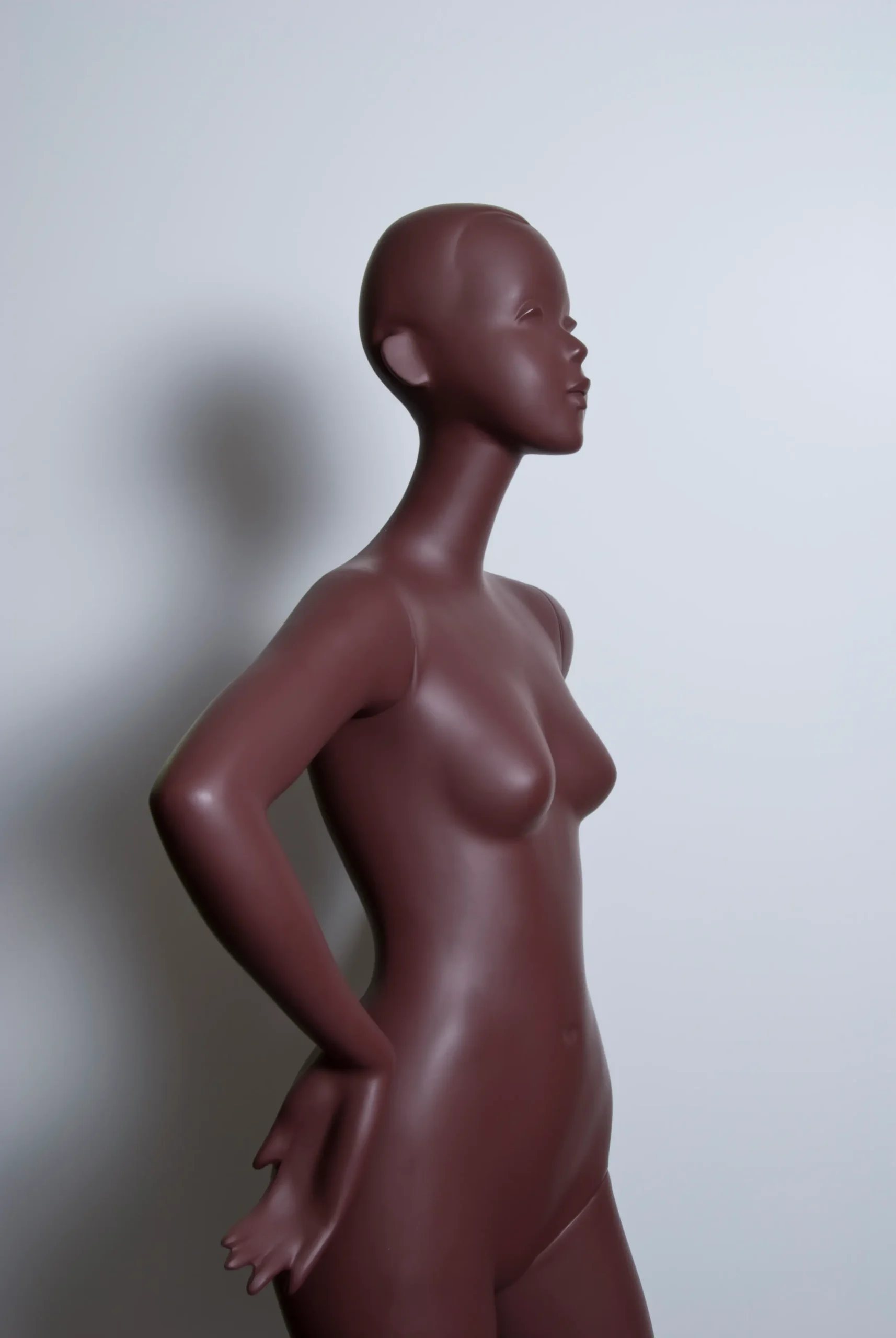 Profile view of a smooth, dark brown female mannequin with hands on hips against a white background.