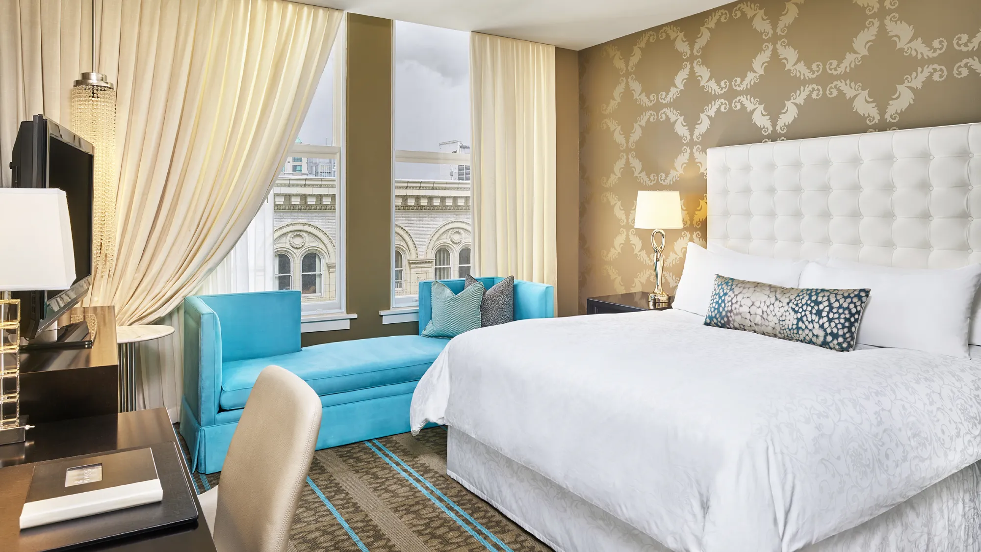 Luxury hotel room featuring a white tufted bed, teal chaise lounge, and patterned wallpaper.